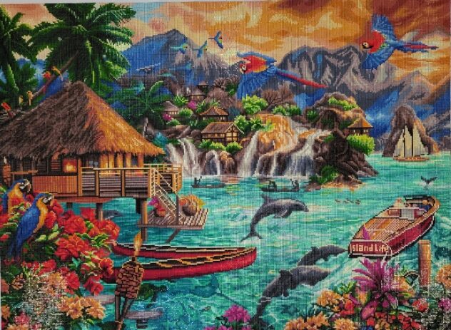 This is a photo of the finished diamond painting of Island Life. It is a picture if a hut near the water with foliage, flowers, and bird around. There are dolphins jumping out of the water and several boats in the water. f